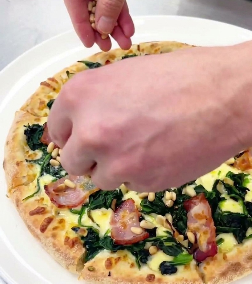 Eine Hand streut kleine geröstete Pinienkerne über eine frisch geschnittene Pizza auf einem weißen Teller.