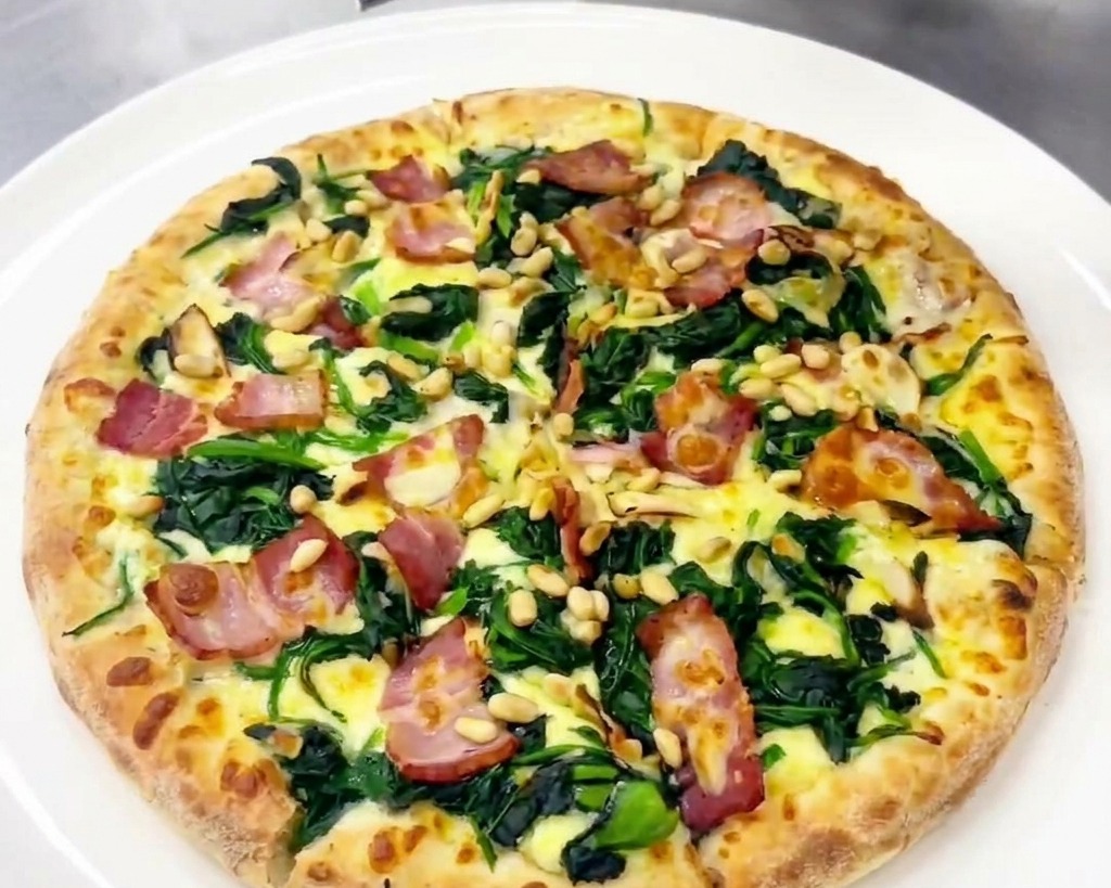 Eine Person hobelt frische Späne eines Hartkäses über eine fertig angerichtete Pizza mit Spinat und Speck.