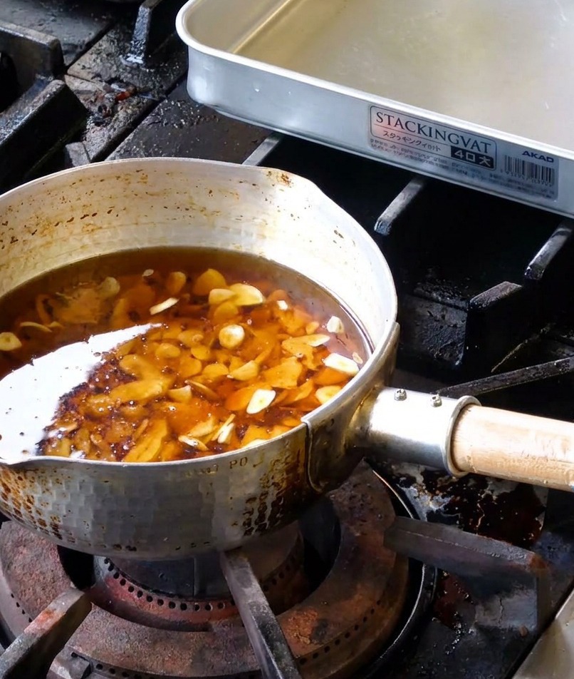 Verschiedene rohe Knoblauchscheiben in einer Mischung aus flüssigem Schmalz und Sesamöl in einem Metalltopf.