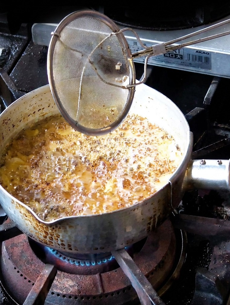 Eine Hand entnimmt mittelbraun frittierte Knoblauchscheiben mit einem Sieb aus dem dunklen Öl.