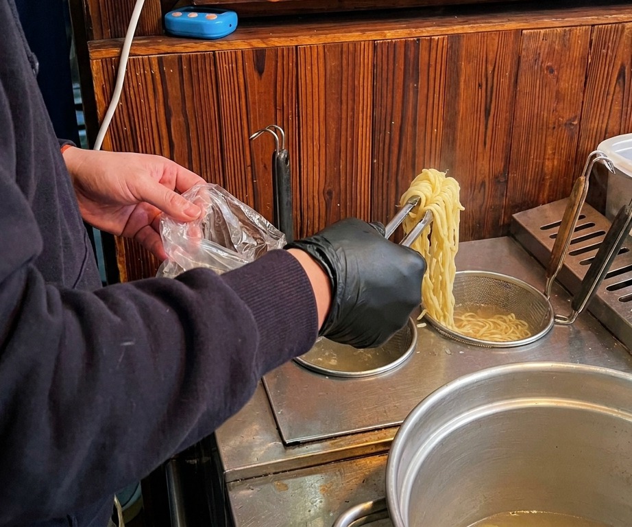 Eine Person mit schwarzen Handschuhen hebt lange, gelbe Ramen-Nudeln mit einer Zange aus kochendem Wasser.