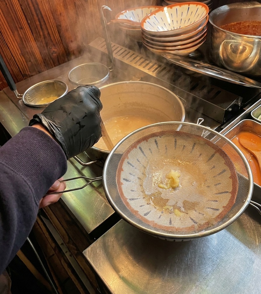Dampfende, cremeweiße Knochenbrühe wird durch ein Metallsieb in eine dekorierte Ramen-Schale gegossen.