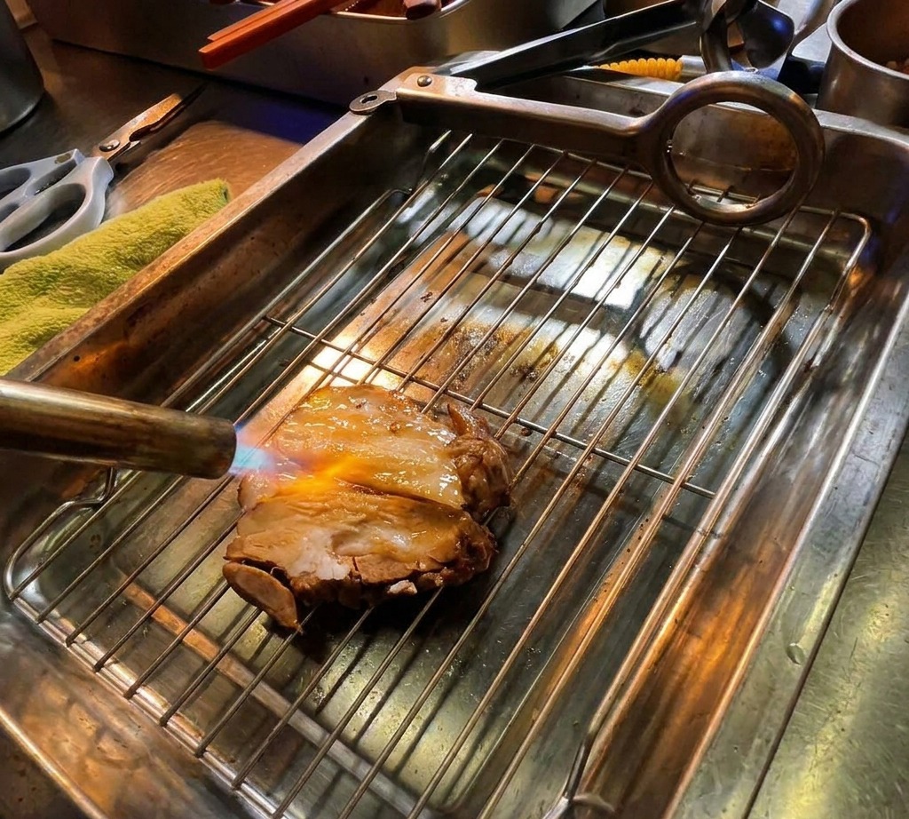 Ein Küchenbrenner flambiert dicke Scheiben von mariniertem Schweine-Chashu auf einem Metallgitter.