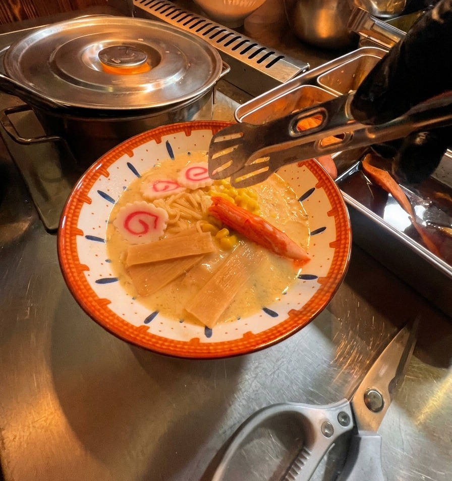 Eine Zange platziert bunte Beilagen wie Fischkuchen, Surimi und Mais in einer Ramen-Schüssel.