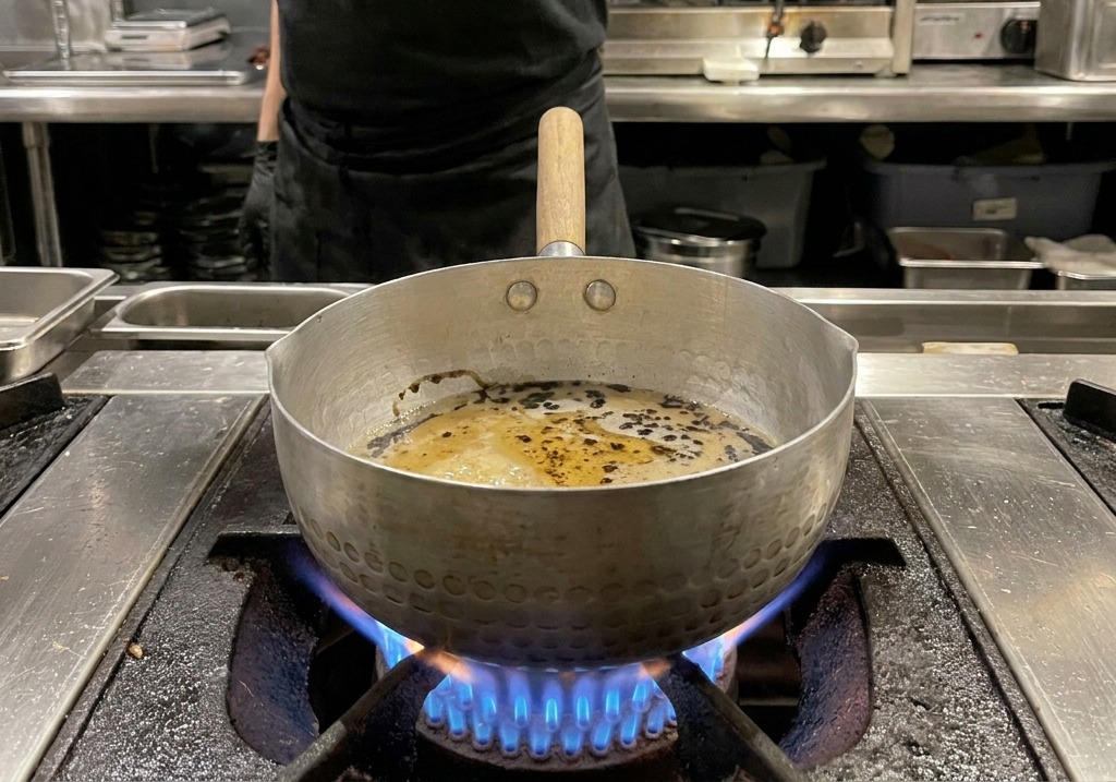 Tiefschwarze Öltröpfchen wirbeln auf der Oberfläche einer hellbraunen Sauce im Topf.