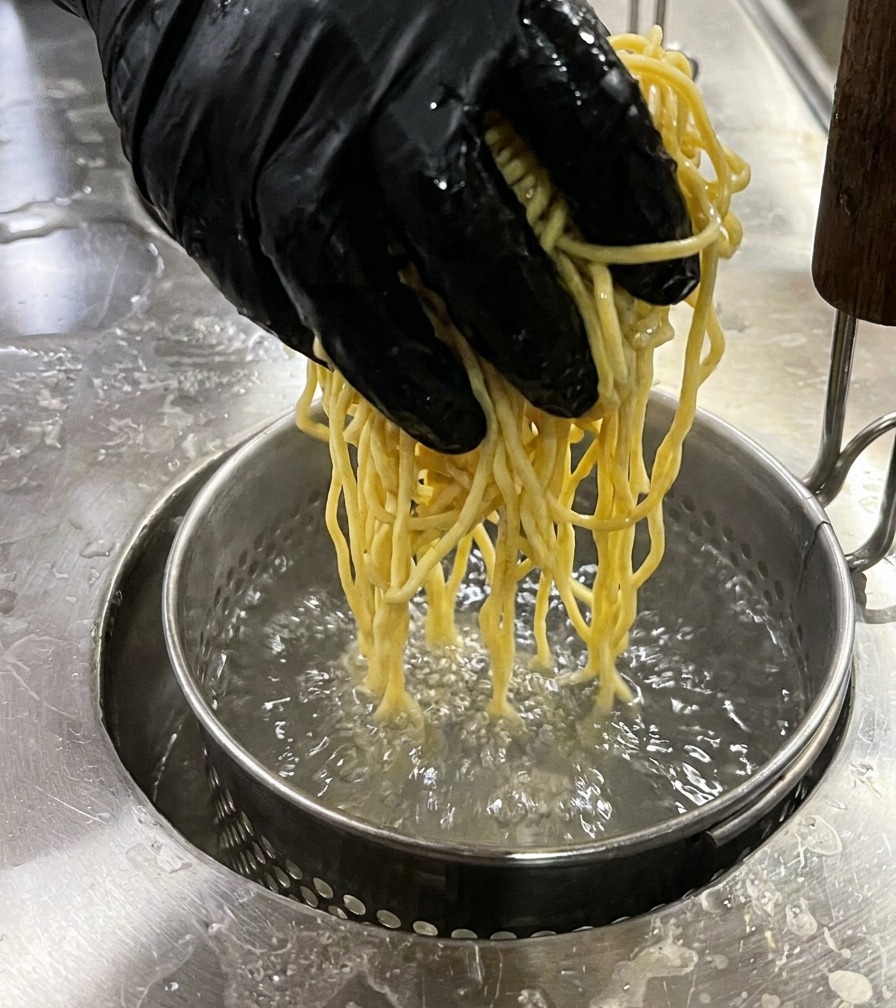Ein Nudelsieb mit gelben Ramen-Nudeln wird in kochendes Wasser abgesenkt.