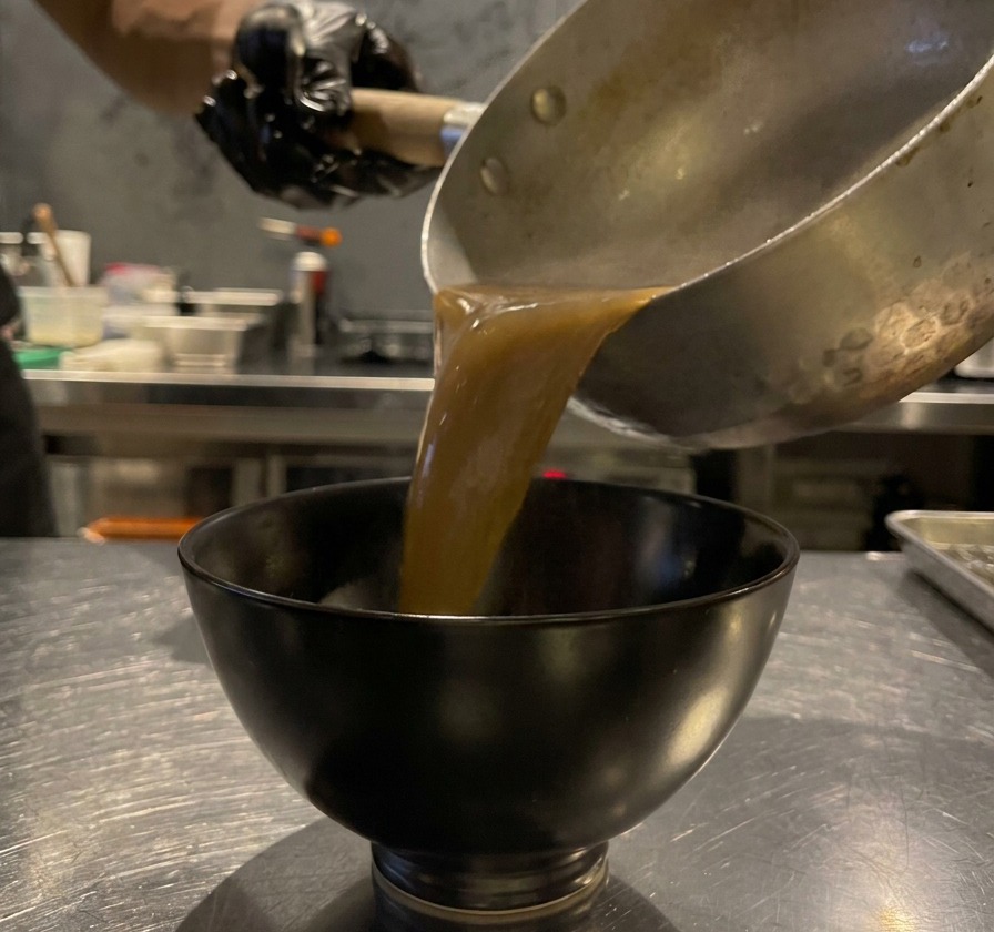 Dicke, braune Knoblauchsauce wird aus einem Topf in eine kleine schwarze Schale gegossen.