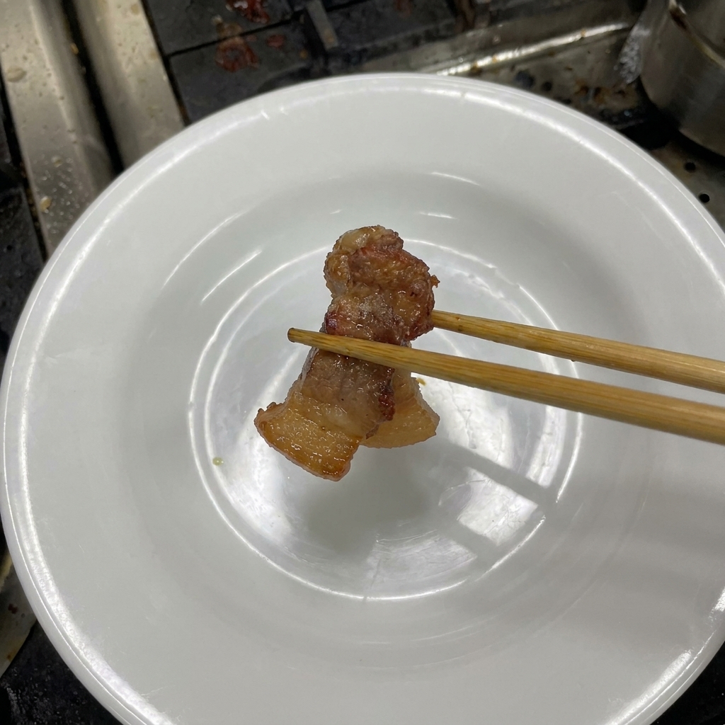 Holzstäbchen halten ein zartes Stück dunkelbraun geschmorten Schweinebauch über einen weißen Teller.