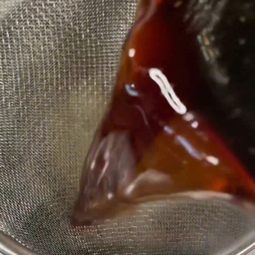 Nahaufnahme einer dunklen, klaren Sauce, die durch ein feinmaschiges Metallsieb gegossen wird.