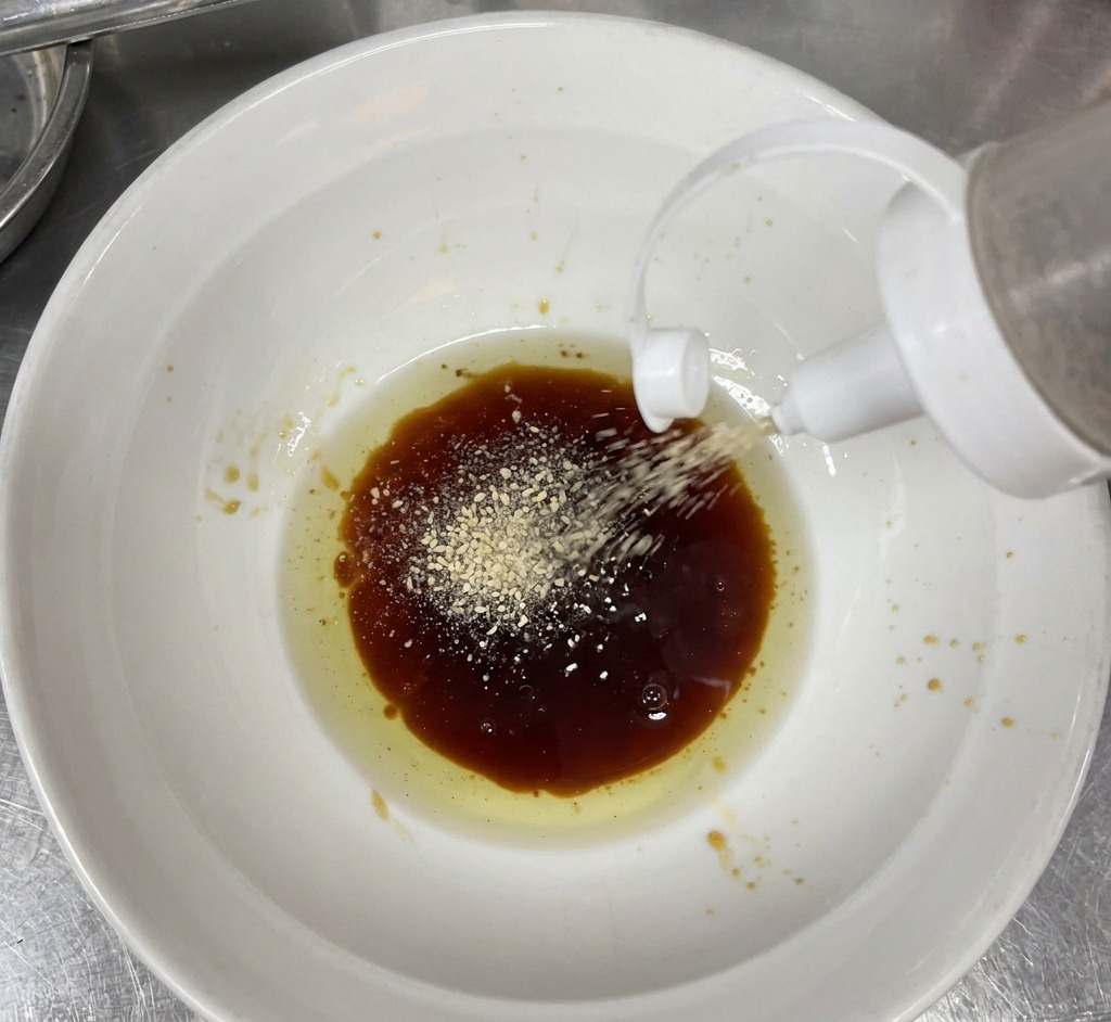 Weißes Sesampulver wird aus einer Flasche in eine Schüssel mit dunkler Sauce und Aromaöl gestreut.