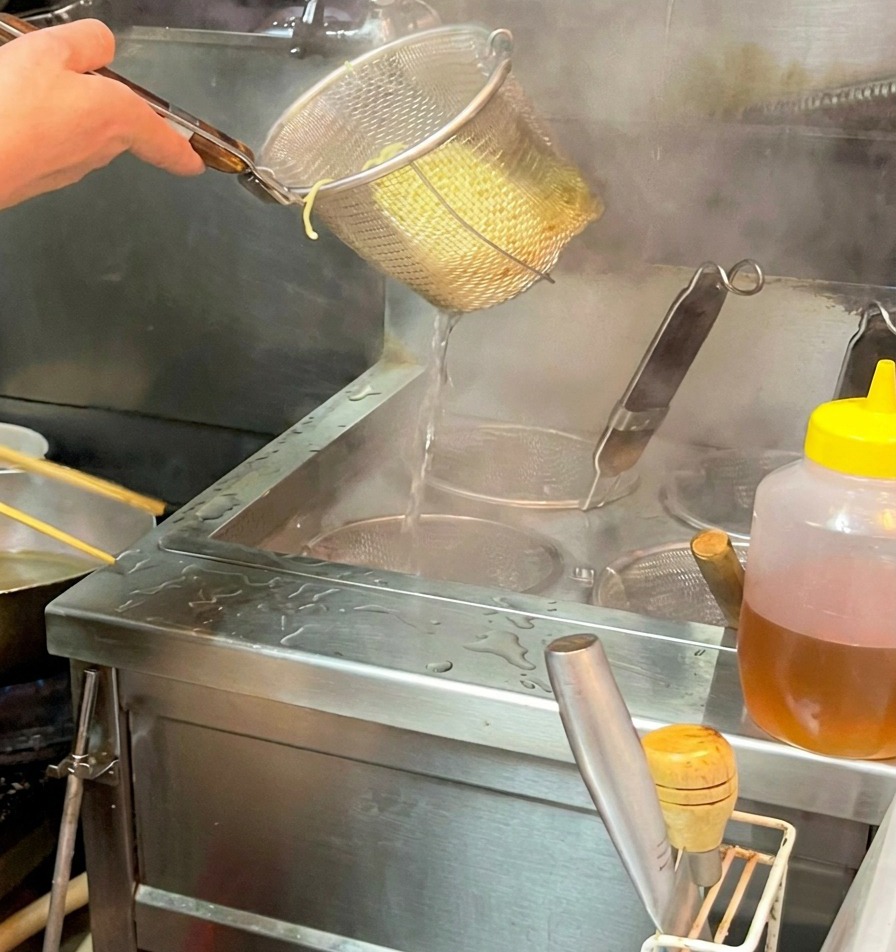 Gekochte Ramen-Nudeln werden aus dem Sieb in die dampfende dunkle Brühe gegeben.