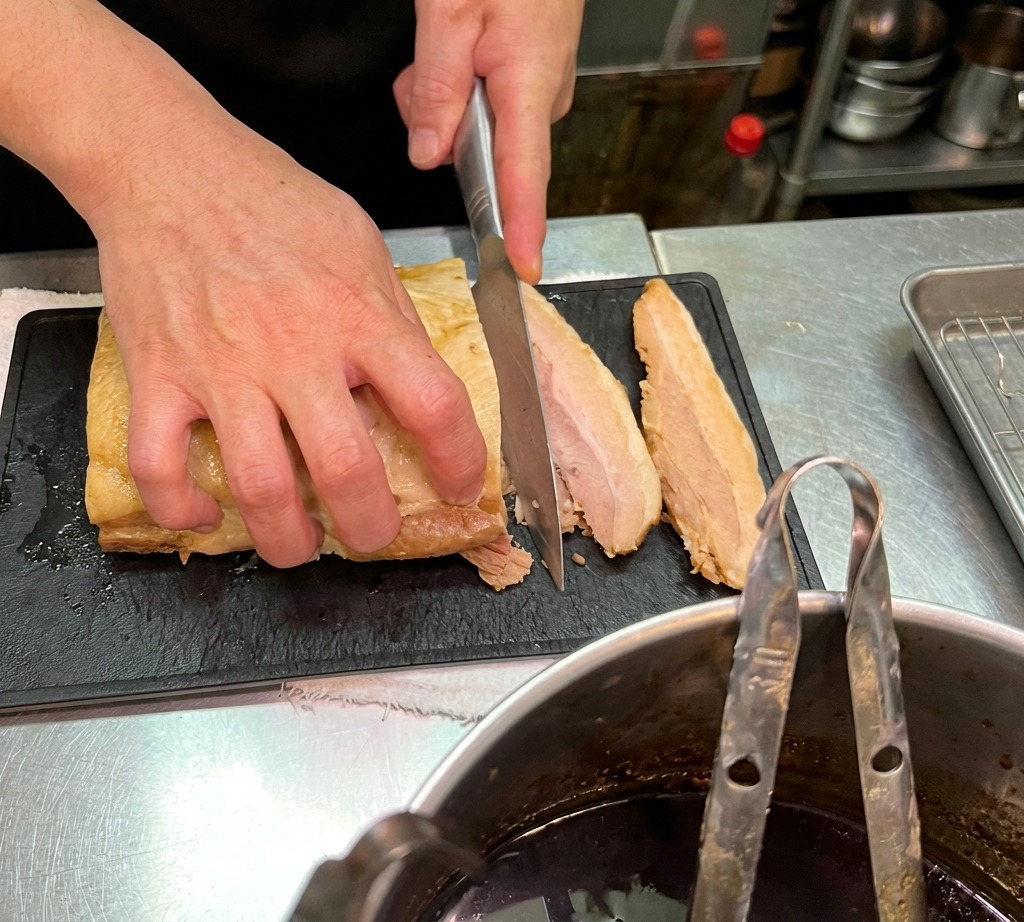 Nahaufnahme eines Kochs, der eine Scheibe zarten Schweinebauch auf einem schwarzen Schneidebrett schneidet.