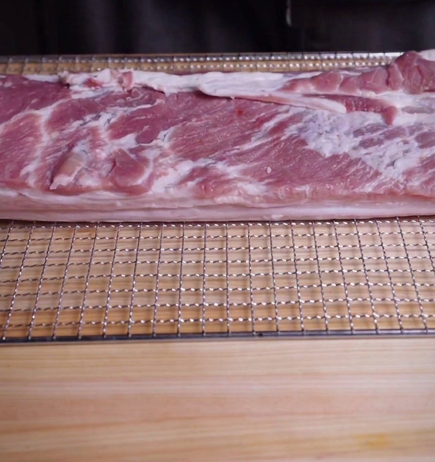 Ein großes Stück roher Schweinebauch auf einem Kuchengitter über einem Holzschneidebrett.