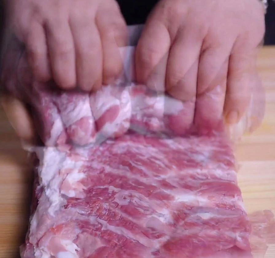 Hände, die ein Stück rohen Schweinebauch auf einem Holzbrett fest zu einem Zylinder rollen.