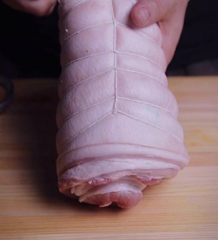 Hände fixieren eine Schweinebauchrolle mit weißem Küchengarn auf einem Holzbrett.