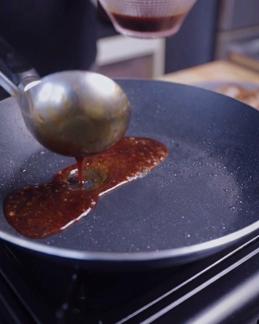 Eine Schöpfkelle gießt eine dunkle, glänzende Sauce über angebratene Schweinebauchscheiben in einer Pfanne.