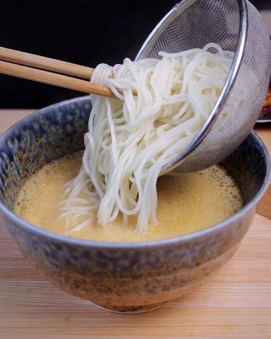 Ein Edelstahlsieb gibt lange weiße Nudeln in eine blaue Schale mit gelber Ramen-Brühe.