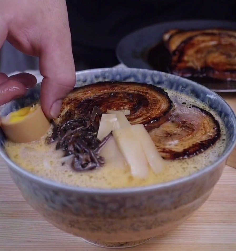 Eine Hand legt ein halbiertes Ei in eine Schale Ramen, die bereits mit Fleisch, Pilzen und Bambus garniert ist.