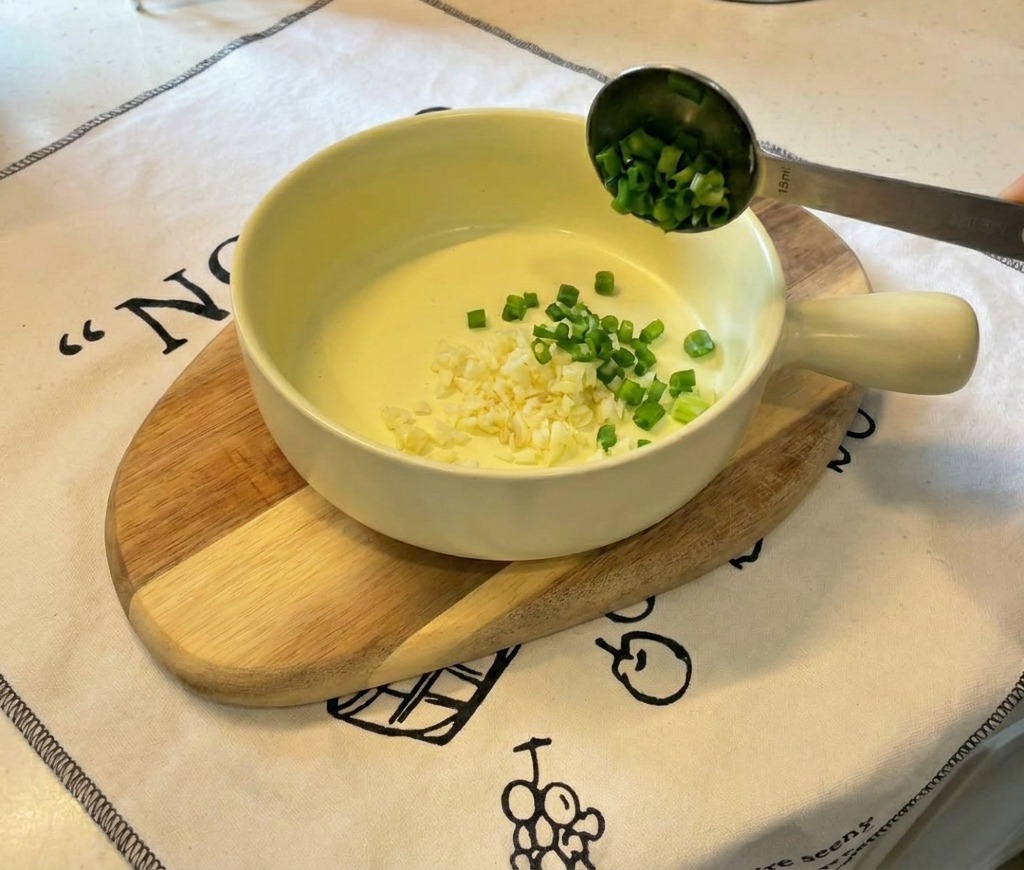 Eine kleine gelbe Keramikschüssel auf einem Holzbrett mit fein gehacktem Knoblauch und geschnittenen Frühlingszwiebeln.