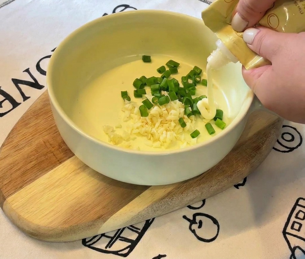 Hände drücken weiße Mayonnaise aus einer gelben Tube in eine Schüssel mit Knoblauch und Frühlingszwiebeln.