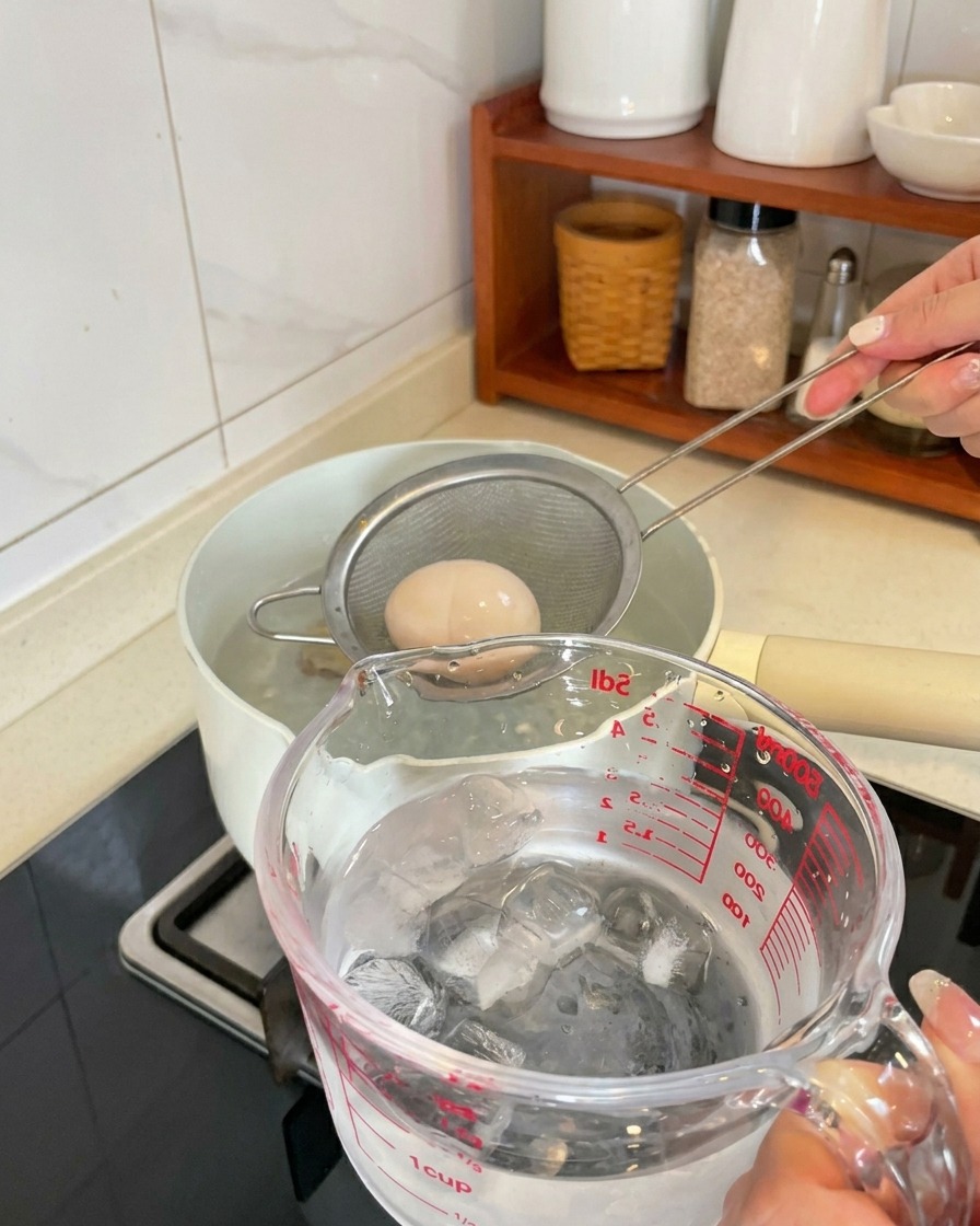 Ein gekochtes Ei wird mit einer Schaumkelle aus dem Topf gehoben und in einen Becher mit Eiswürfeln und Wasser gelegt.