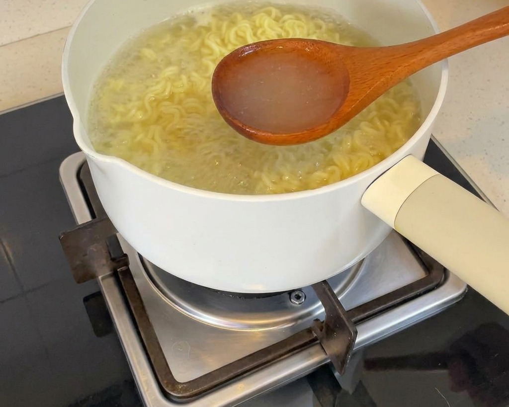 Eine Schöpfkelle hebt klares, dampfendes Nudelwasser aus einem weißen Topf über einer Herdplatte.