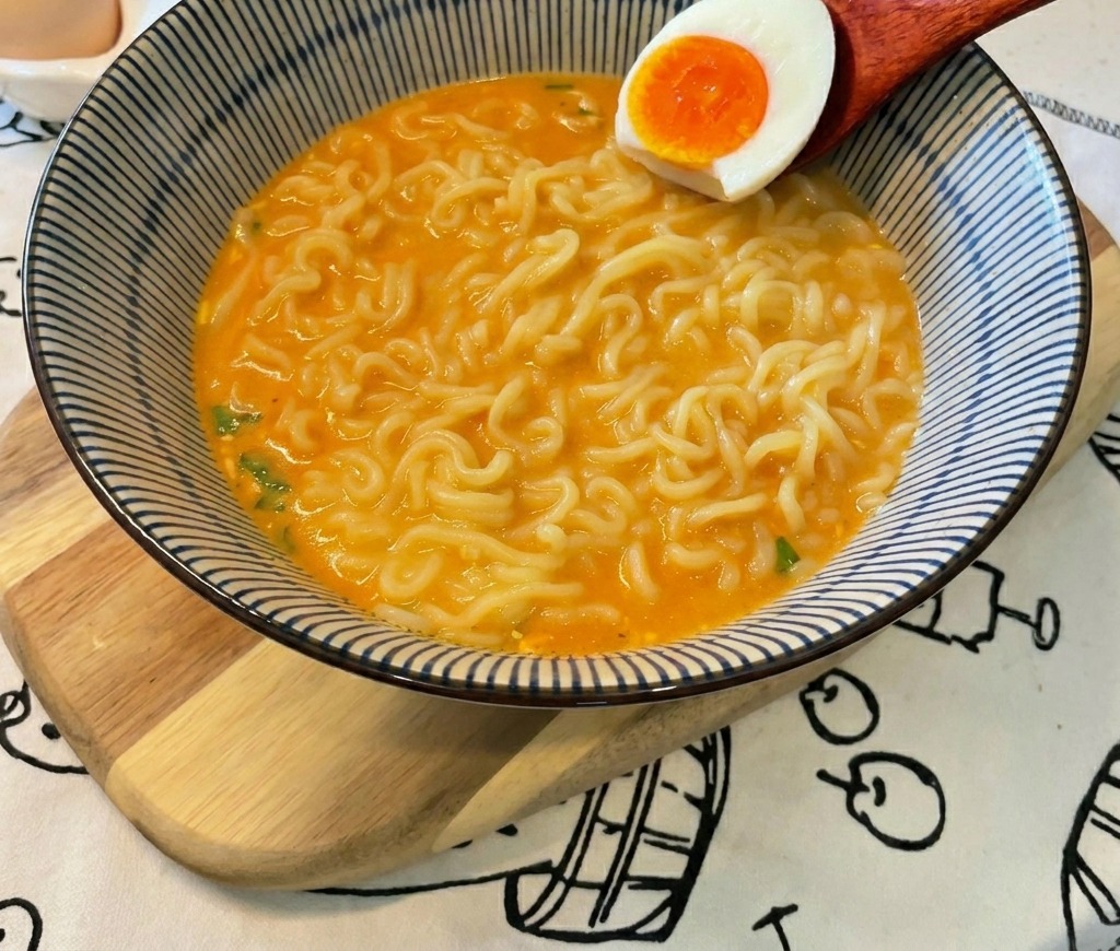 Ein Holzlöffel legt eine halbe Eihälfte mit leuchtend orangefarbenem Dotter in eine Schüssel mit Ramen und Brühe.
