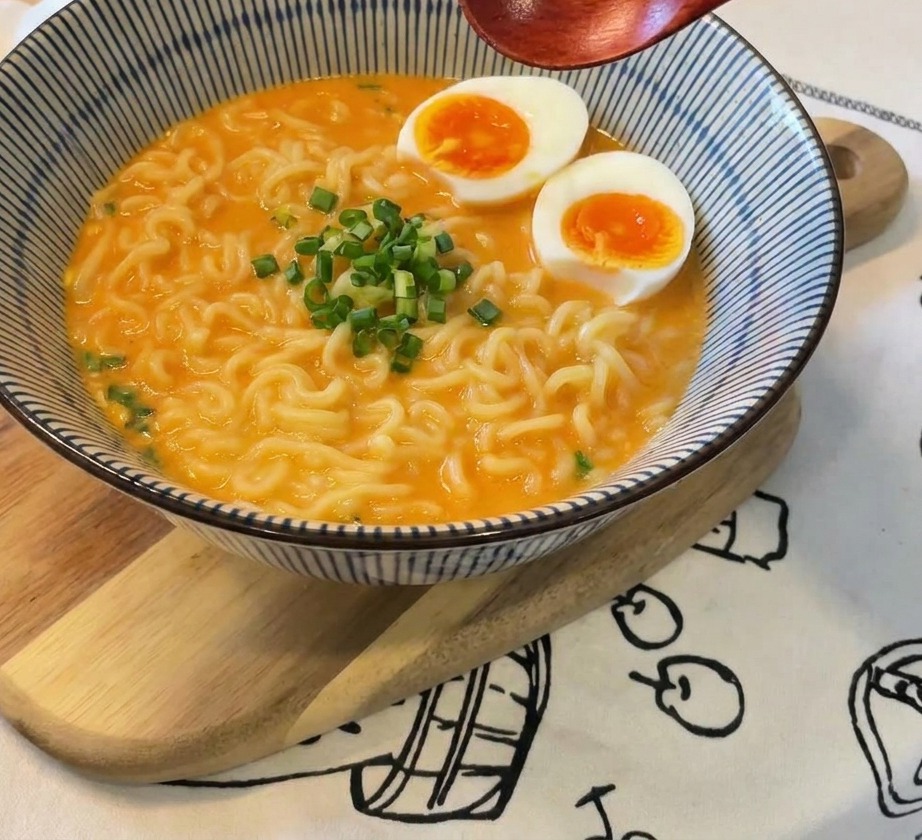 Eine Schüssel Ramen garniert mit Eiern und frischen Frühlingszwiebeln in der Mitte, ein Holzlöffel wird über das Gericht gehalten.