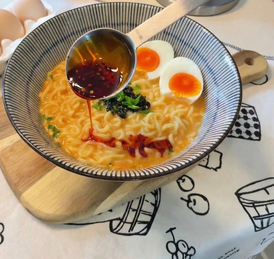 Leuchtend rotes Chiliöl wird aus einem Metalllöffel über die fertige Schüssel Ramen mit Eiern und Zwiebeln gegossen.