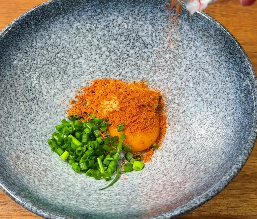Rotes Ramen-Gewürz wird in eine blaue Schüssel mit einem Eigelb und Frühlingszwiebeln geschüttet.