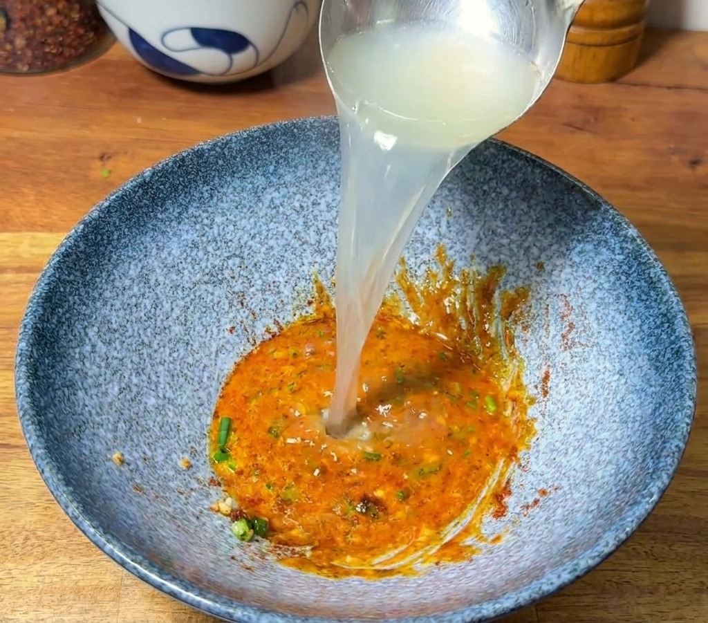 Eine Schöpfkelle gießt dampfendes Wasser in die Schüssel mit der Würzpaste, wodurch eine cremige Brühe entsteht.