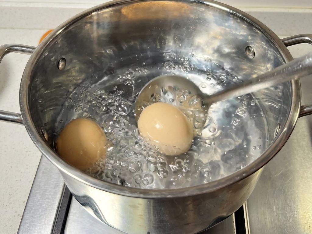 Ein Schaumlöffel hebt zwei Eier aus einem Topf mit kochendem Wasser.