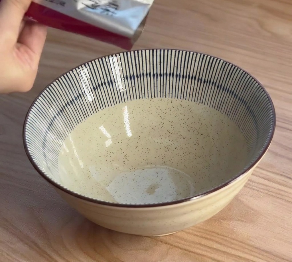 Eine Hand drückt dunkle Miso-Paste aus einer Folienpackung in eine blau-weiß gestreifte Schüssel.