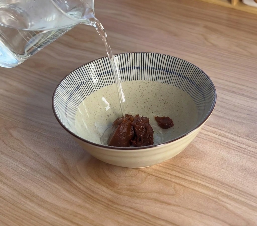 Dampf steigt auf, während kochendes Wasser aus einem Messbecher auf die Misopaste gegossen wird.