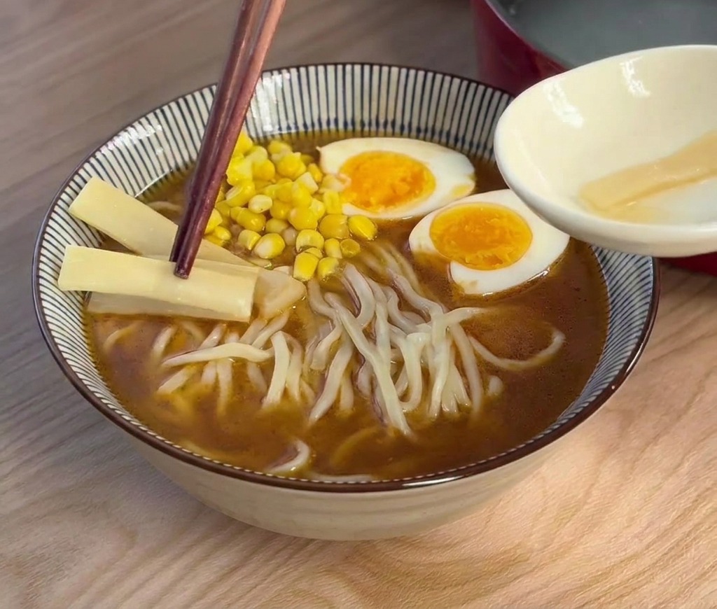 Holzstäbchen legen helle Bambussprossen in die Ramen-Schüssel neben Ei und Mais.