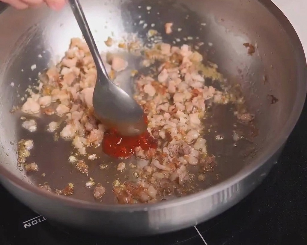 Ein Metalllöffel gibt eine Portion rote scharfe Bohnenpaste zu dem angebratenen Fleisch in der Pfanne.