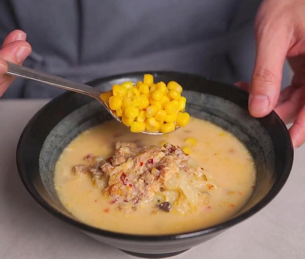 Ein Metalllöffel gibt eine Portion goldenen Mais auf die Oberfläche der Ramen-Suppe.