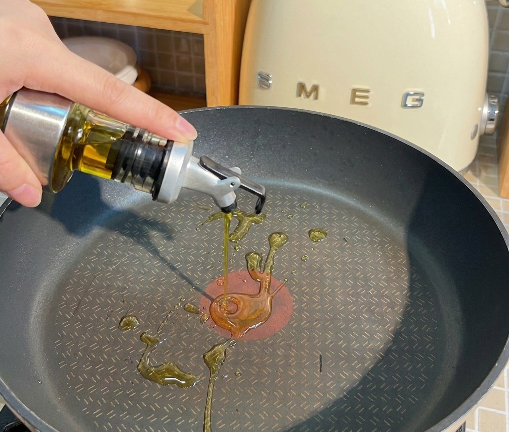 Speiseöl wird aus einem Glasbehälter in eine schwarze, beschichtete Pfanne gegossen.