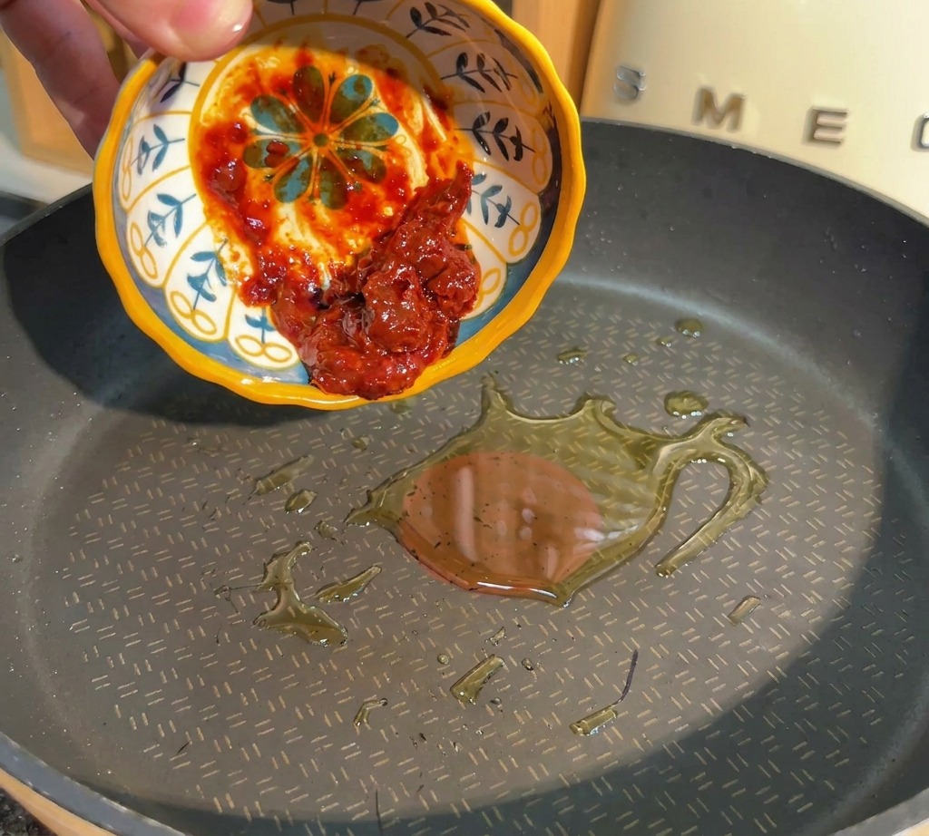Eine Hand gibt scharfe rote Bohnenpaste aus einer kleinen Schale in eine Pfanne mit heißem Öl.