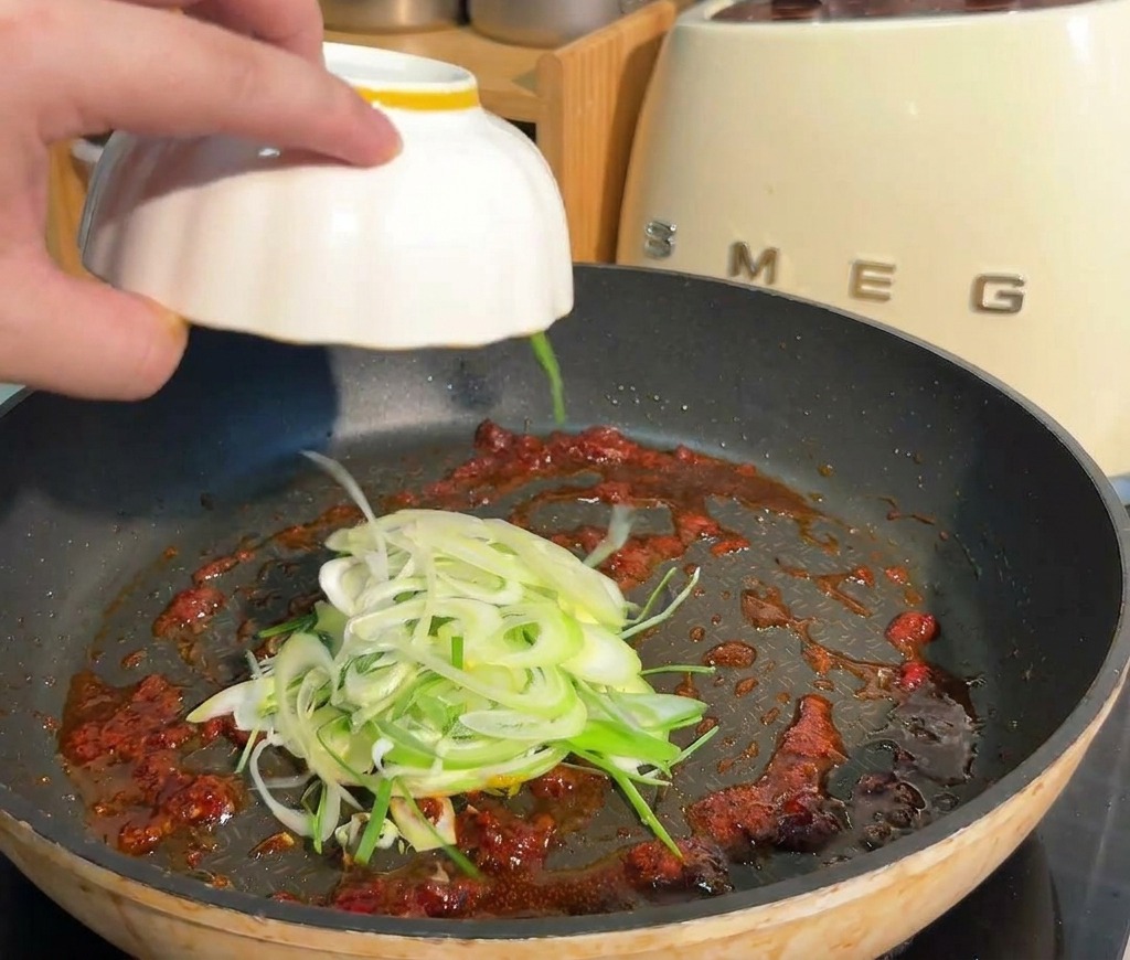 Geschnittene Frühlingszwiebeln werden aus einer weißen Schale in eine Pfanne mit roter Saucenbasis gegeben.