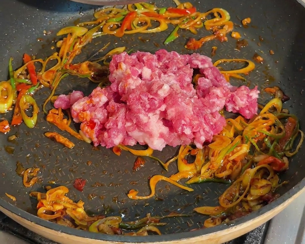 Rohes Schweinehackfleisch wird in eine Pfanne mit angebratenen Frühlingszwiebeln und roter Bohnenpaste gegeben.