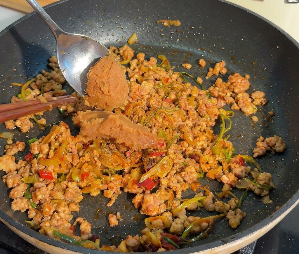 Ein Metalllöffel gibt eine Portion dicke braune Misopaste in eine Pfanne mit gegartem Hackfleisch.