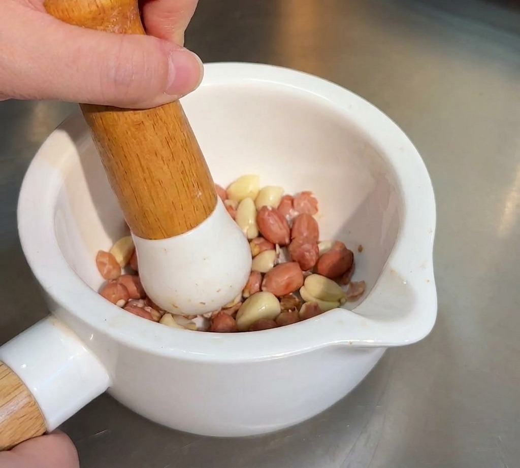 Eine Person zerstößt geröstete Erdnüsse und Sesam mit einem Holzstößel in einem Keramikmörser.
