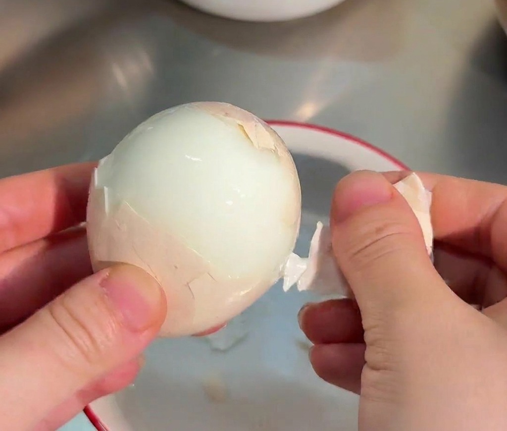 Hände pellen vorsichtig die Schale von einem glatten, weißen weichgekochten Ei über einer kleinen Schale ab.