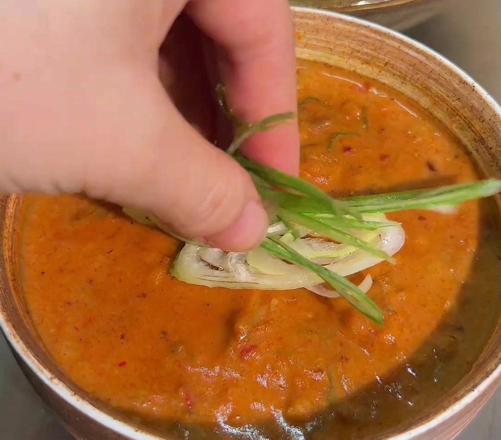 Eine Hand platziert fein geschnittene Frühlingszwiebelstreifen in der Mitte einer orangefarbenen Dipsauce.