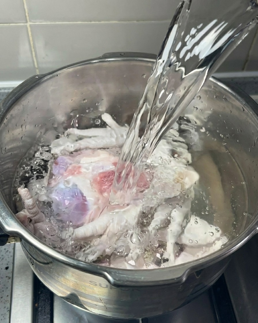 Ein Strahl klaren Wassers wird in einen Edelstahltopf mit rohen Schweineknochen und Hühnerfüßen gegossen.