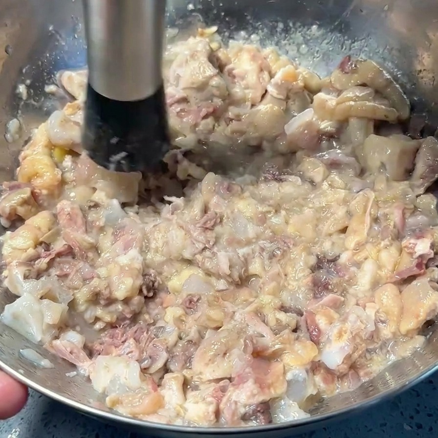 Ein Kartoffelstampfer zerdrückt gegartes Fleisch zu einer groben Paste in einer Edelstahlschüssel.