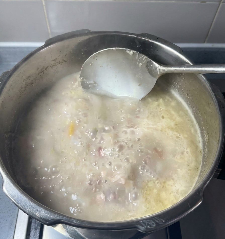 Eine Schöpfkelle rührt in einer cremigen, milchig-weißen Knochenbrühe mit sichtbaren Fleischstücken.