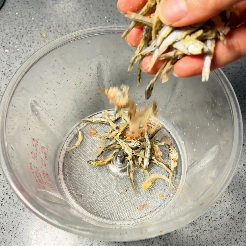 Eine Hand schüttet getrocknete Sardellen und Bonitoflocken in einen Standmixer.