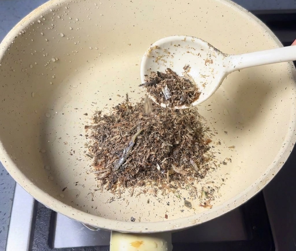Ein Löffel mit braunem Fischpulver wird in einen kleinen Stieltopf gegeben.